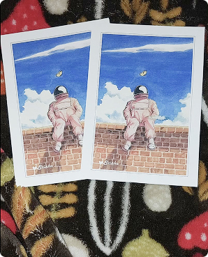 Two astronaut-on-a-brick-wall postcards lying on dark floral fabric