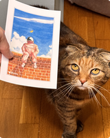 Astronaut-on-a-brick-wall postcard held up beside a curious tabby cat