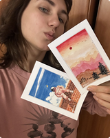 Two watercolor postcards held up: a pink desert sunset and an astronaut on a brick wall