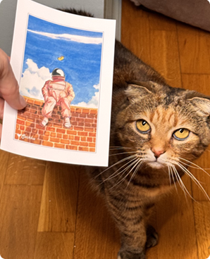 Astronaut-on-a-brick-wall postcard held up beside a curious tabby cat