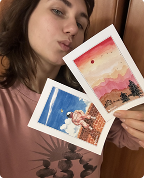 Two watercolor postcards held up: a pink desert sunset and an astronaut on a brick wall