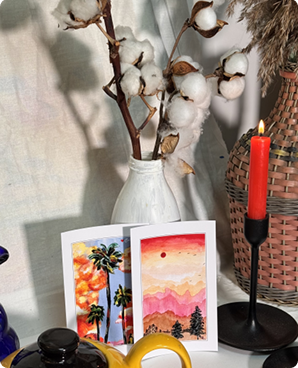 Two palm-tree sunset postcards propped near dried cotton stems and a red candle