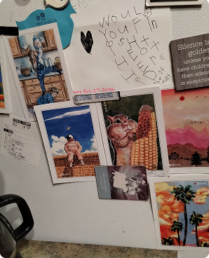 A fridge door covered with magnets and quirky art postcards including a winged cat
